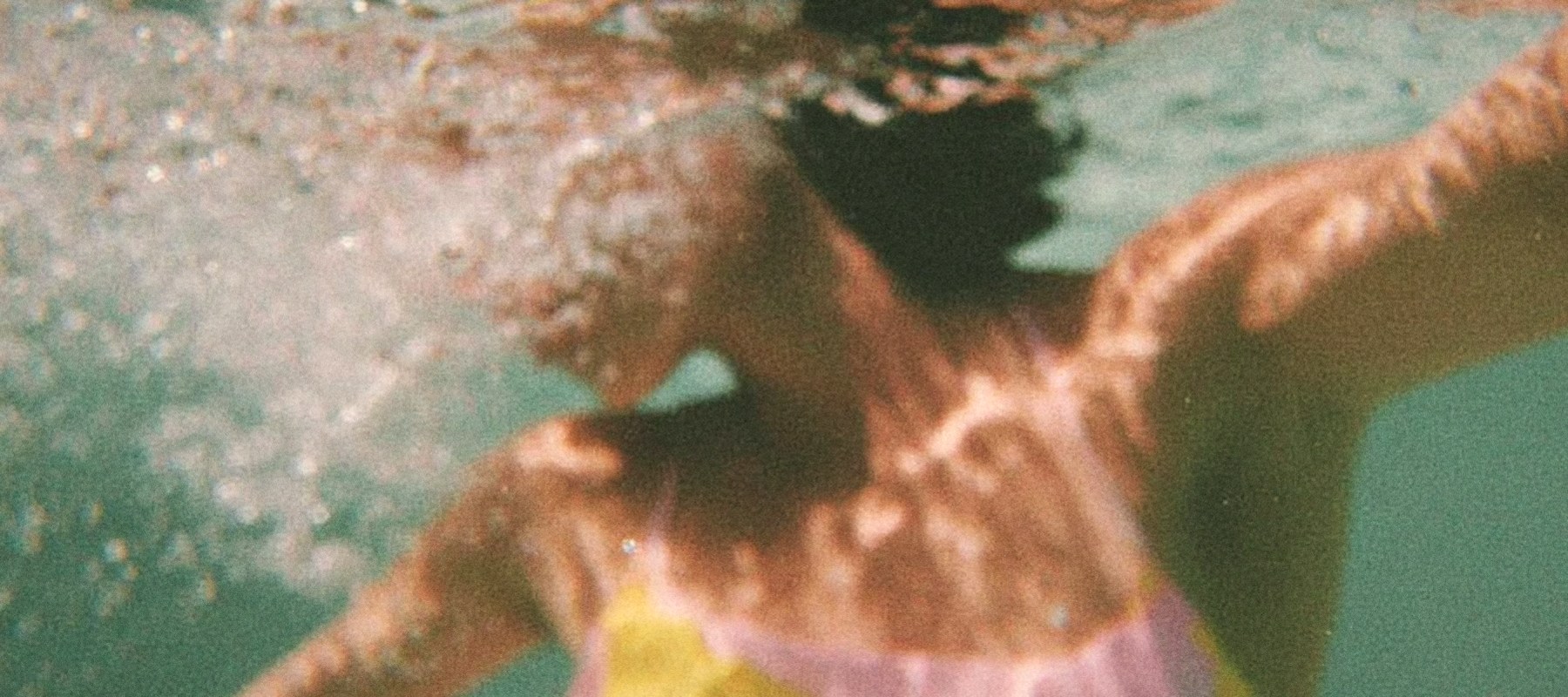 A swimmer underwater in turquoise water, pink and yellow swimsuit, film photograph
